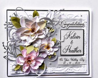 May include: A white card with a silver embossed design and a floral embellishment. The card says "Congratulations Kelvin & Heather On Your Wedding Day 01.16.2023".