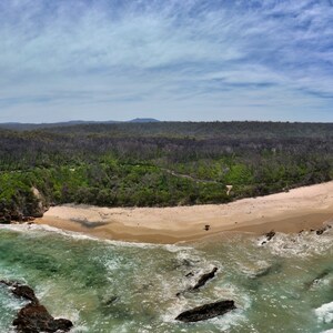 Puede incluir: Vista a&eacute;rea panor&aacute;mica de una costa con playa de arena, agua turquesa y formaciones rocosas. Un denso bosque bordea la playa, y el cielo es azul con nubes dispersas. Las olas del oc&eacute;ano rompen en la orilla.