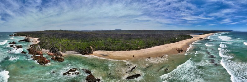 Puede incluir: Vista a&eacute;rea panor&aacute;mica de una costa con playa de arena, agua turquesa y formaciones rocosas. Un denso bosque bordea la playa, y el cielo es azul con nubes dispersas. Las olas del oc&eacute;ano rompen en la orilla.