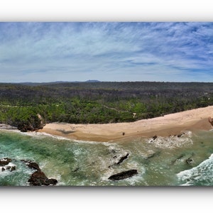 Puede incluir: Vista a&eacute;rea panor&aacute;mica de una playa con agua turquesa, olas blancas y arena. Afloramientos rocosos y un bosque verde bordean la costa bajo un cielo azul con nubes dispersas. La imagen es un rect&aacute;ngulo horizontal largo.