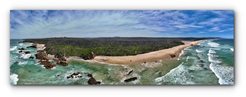 Puede incluir: Vista a&eacute;rea panor&aacute;mica de una playa con agua turquesa, olas blancas y arena. Afloramientos rocosos y un bosque verde bordean la costa bajo un cielo azul con nubes dispersas. La imagen es un rect&aacute;ngulo horizontal largo.