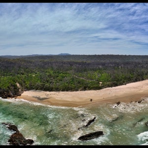 Puede incluir: Vista a&eacute;rea panor&aacute;mica de una costa con playa de arena, agua turquesa y formaciones rocosas. Un denso bosque separa la playa de una laguna. El cielo es azul con nubes dispersas, creando un paisaje pintoresco.