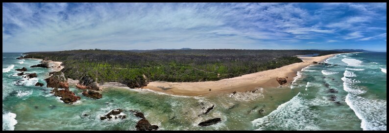 Puede incluir: Vista a&eacute;rea panor&aacute;mica de una costa con playa de arena, agua turquesa y formaciones rocosas. Un denso bosque separa la playa de una laguna. El cielo es azul con nubes dispersas, creando un paisaje pintoresco.