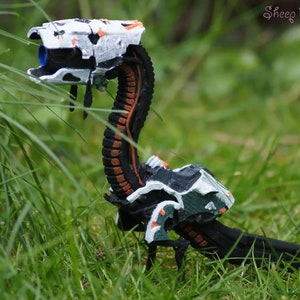 Könnte beinhalten: Ein 3D-gedrucktes Modell einer schwarzen, weißen und orangefarbenen Roboterkreatur mit langem Hals und einem Kopf, der einem Gewehr ähnelt. Die Kreatur ist in einer Gehposition mit nach unten geneigtem Kopf dargestellt. Das Modell steht auf einem Bett aus grünem Gras.