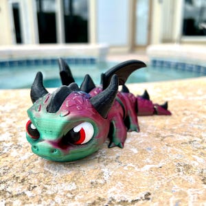 May include: A small, green and purple dragon figurine with black horns and wings. The dragon has large, red eyes and a textured surface.