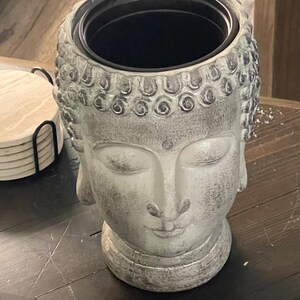 May include: A grey Buddha head planter with a black interior. The planter features a serene expression with closed eyes and a detailed hairstyle. The surface has a weathered, textured appearance, suggesting a stone or concrete material. The planter is on a dark wooden surface.