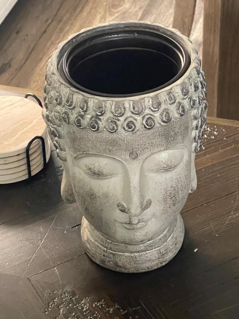 May include: A grey Buddha head planter with a black interior. The planter features a serene expression with closed eyes and a detailed hairstyle. The surface has a weathered, textured appearance, suggesting a stone or concrete material. The planter is on a dark wooden surface.