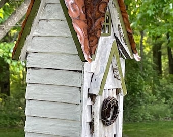 Peut inclure: Un nichoir en bois blanc rustique avec un toit en cuivre décoratif à motifs et une bordure verte. Le nichoir comprend une petite fenêtre, une couronne de brindilles et un perchoir en bois. Le nichoir est suspendu à une branche d'arbre.
