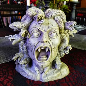 May include: A detailed bust of Medusa with snakes for hair, rendered in shades of gray and green. The sculpture features an open mouth with visible fangs and a cracked, aged appearance, evoking a sense of ancient mythology and horror.