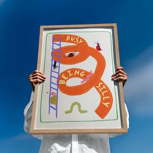 Könnte beinhalten: Ein gerahmter rechteckiger Druck mit einer geschwungenen orangefarbenen Rutsche, auf der der Text BUSY BEING SILLY entlang der Kurve steht. Die Illustration zeigt eine blaue Leiter, einen Marienkäfer, eine Schnecke, einen Wurm und eine grüne Raupe.