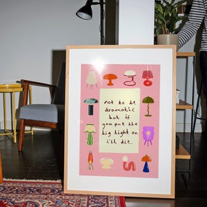 May include: A framed rectangular print with a pink background featuring various colorful lamp illustrations surrounding a central pale yellow rectangle containing the black handwritten text: not to be dramatic but if you put the big light on i'll die.