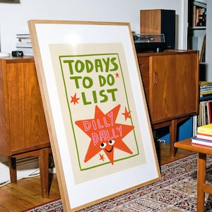 May include: A framed wall art print featuring a cream background with a green rectangular border. The text reads TODAYS TO DO LIST in green. Below, a large orange star shape with a smiling face and eyes contains the pink text DILLY DALLY.