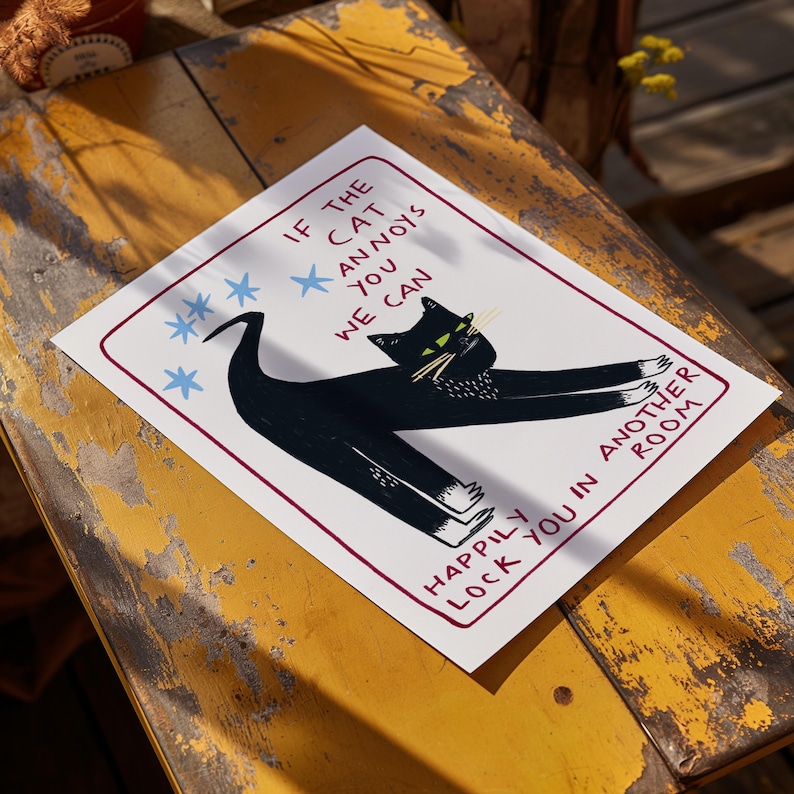 以下が含まれることがあります： 黒猫のイラストと「If the cat annoys you we can happily lock you in another room」というテキストがプリントされた白いプリント。猫は緑色の目と白い足を持っています。青い星のイラストも含まれています。