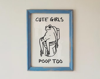 May include: A framed art print with a blue distressed wooden frame. The black and white illustration depicts a frog sitting on a toilet with the text "CUTE GIRLS POOP TOO". The print is a humorous bathroom decor item.