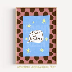 May include: A framed art print with a pink background featuring brown oval shapes. The center has a blue rectangle with yellow stars and the phrase "grubs up bitches" in a white speech bubble. The print is in a wooden frame.