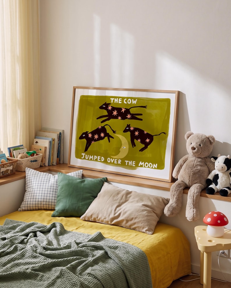 May include: A framed rectangular wall art print featuring a dark olive green background with three black cow silhouettes decorated with pink stars, a yellow crescent moon, and the white text THE COW JUMPED OVER THE MOON.