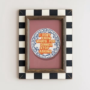 May include: A framed print with a black and white chequered border. The print features a round plate design with the text "I DON'T KNOW WHAT'S FOR DINNER STOP ASKING" in orange letters. The background is a muted pink.