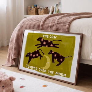 May include: A framed rectangular wall art print featuring three dark brown cows with pink star patterns jumping over a yellow crescent moon on an olive green background, with the text THE COW JUMPED OVER THE MOON printed in white.