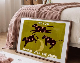 Puede incluir: Una lámina de arte mural rectangular enmarcada que muestra tres vacas de color marrón oscuro con patrones de estrellas rosas saltando sobre una luna creciente amarilla sobre un fondo verde oliva, con el texto THE COW JUMPED OVER THE MOON en blanco.