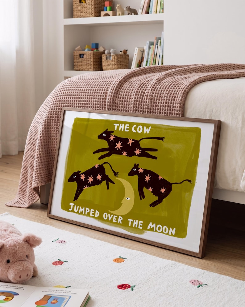 May include: A framed rectangular wall art print featuring three dark brown cows with pink star patterns jumping over a yellow crescent moon on an olive green background, with the text THE COW JUMPED OVER THE MOON printed in white.