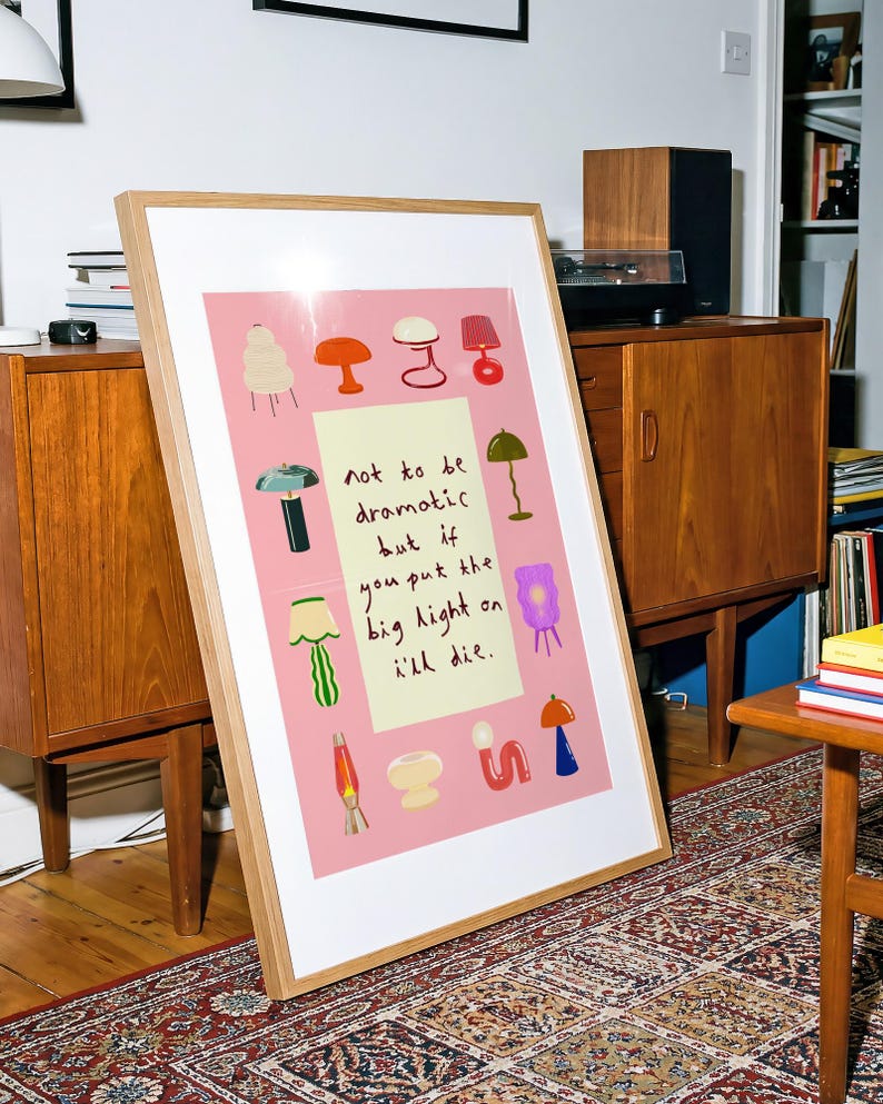 May include: A framed rectangular art print with a pink background featuring various colorful lamp illustrations surrounding a central cream-colored rectangle with the handwritten text: not to be dramatic but if you put the big light on i'll die.
