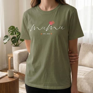 Puede incluir: Camiseta verde oliva con la palabra "mama" en cursiva, un diseño floral rosa y naranja, y el texto "EST. 2026". La camiseta es de cuello redondo y manga corta.