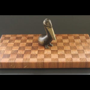 Peut inclure: Une planche à découper en bois avec un motif en damier, faite de bois clair et foncé. Une petite figurine de pélican en métal est posée sur la planche.