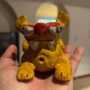 May include: A handmade, yellow ceramic creature figurine with a red nose and white-rimmed eyes. The figurine has visible teeth and is posed on all fours. The creature's ears are upright, and its paws are detailed with claw marks.