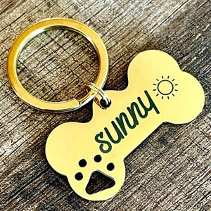 以下が含まれることがあります： 金の骨の形をした犬のタグ。足跡、太陽、そして「Sunny」という名前が刻まれています。タグは金のキーリングに取り付けられています。