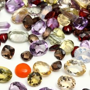 Puede incluir: Una colección de varias piedras preciosas de diferentes formas y tamaños, incluyendo amatista, citrino, cuarzo ahumado y cornalina. Las piedras preciosas están dispuestas sobre un fondo blanco.