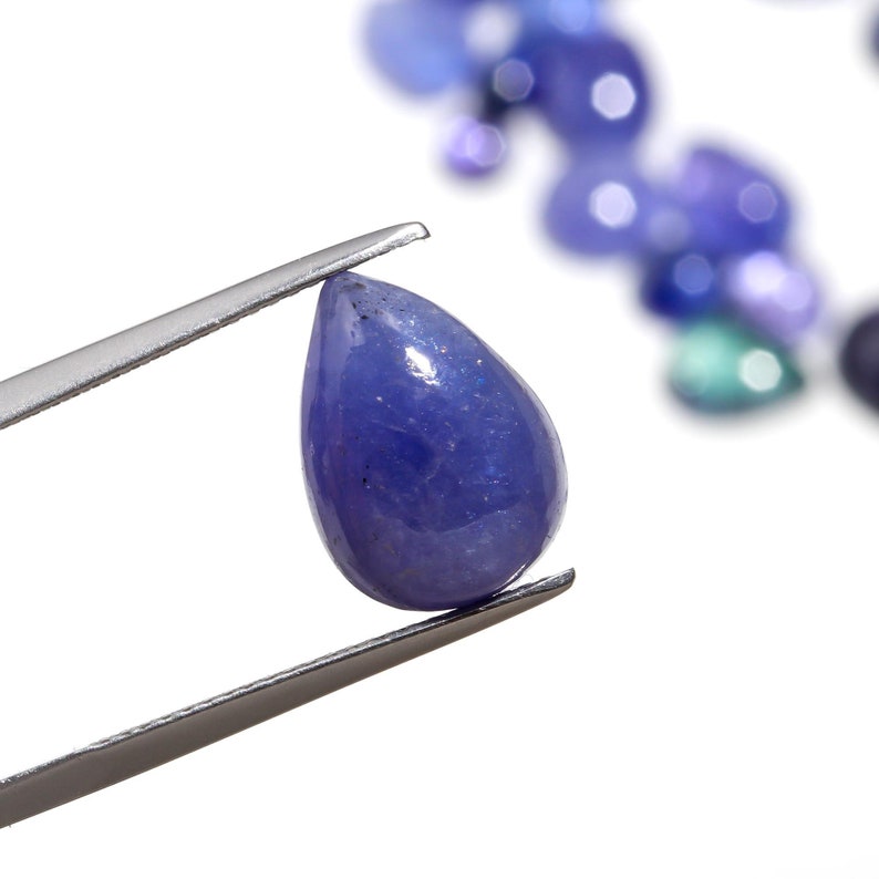 May include: A teardrop-shaped, purple gemstone held by tweezers. The gemstone is faceted and has a shiny, polished finish.