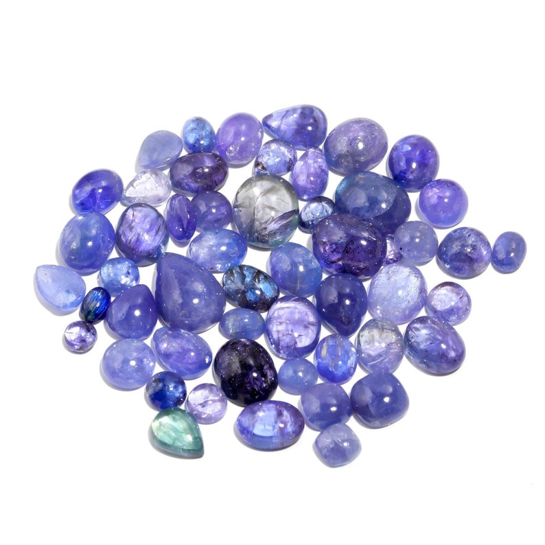 May include: A collection of blue and purple gemstones, likely Tanzanite, in various shapes and sizes. The gemstones are arranged in a loose pile on a white background.