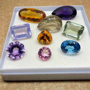 May include: A selection of multi-coloured gemstones in a white tray. The gems are in various shapes, including oval, pear, and rectangular cuts. The colours range from purple and blue to yellow and green.