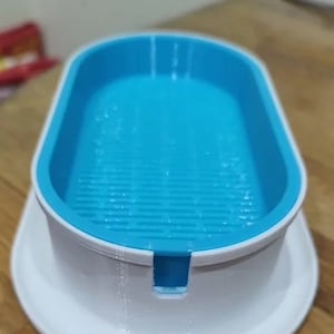 Puede incluir: Un plato de plástico blanco con un inserto de plástico azul. El inserto tiene un patrón de rejilla y una pequeña abertura en el centro.