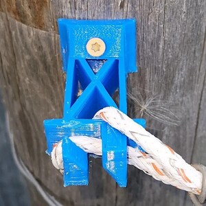 Op de afbeelding: Een blauwe plastic touwklemblok gemonteerd op een houten paal. De klemblok heeft een stervormige schroef die hem op zijn plaats houdt. Een wit touw met oranje accenten is door de klemblok geregen.