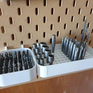 May include: A collection of various drill bits and screwdriver bits organized in white plastic trays. The bits are silver and black, and the trays are mounted on a pegboard. The pegboard is a light brown color.