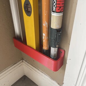 Può includere: Un portaoggetti a parete rosso in plastica che contiene un livello, un'asta di prolunga per rullo per pittura e un'asta di prolunga per pennello per pittura Sherwin-Williams Wooster.