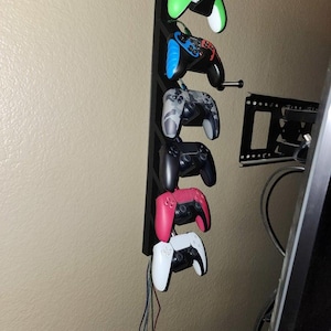 Op de afbeelding: Een zwarte, aan de muur gemonteerde gamecontrollerhouder met zes controllers in verschillende kleuren: groen, blauw, camouflage, zwart, roze en wit. De houder heeft een eenvoudig, verticaal ontwerp.