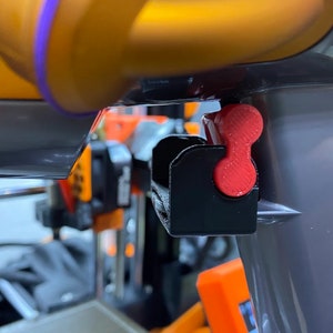 May include: A close-up of a black 3D printer part with a red lever. The lever is attached to a black plastic housing. The lever is in the up position.