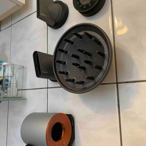 Op de afbeelding: Een set van vier föhndopjes, waaronder een diffuser, een concentratormondstuk, een gladmakende mondstuk en een ronde borstel. De dopjes zijn allemaal in grijstinten en zwart en zijn bevestigd aan een witte betegelde muur.