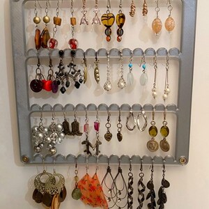 Peut inclure: Un porte-boucles d'oreilles gris avec trois rangées de crochets. Le porte-boucles est rempli d'une variété de boucles d'oreilles, y compris des boucles d'oreilles en argent, en or et des perles colorées.