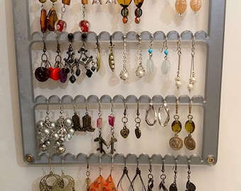 May include: A grey earring holder with three rows of hooks. The holder is filled with a variety of earrings, including silver, gold, and colourful beads.
