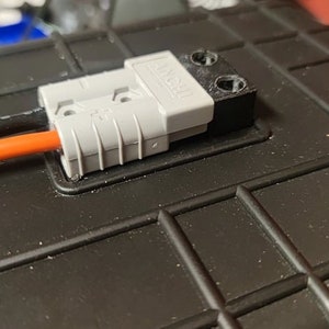 以下が含まれることがあります： グレーとブラックの電気コネクタのクローズアップ。オレンジとブラックのワイヤーが接続されています。コネクタは、テクスチャ加工された表面を持つブラックの四角いボックスに差し込まれています。