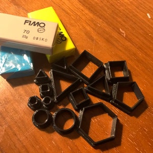 May include: A set of 16 black plastic cookie cutters in various shapes, including circles, squares, triangles, and hexagons. There are also three blocks of polymer clay in the image: one light blue, one beige, and one yellow. The yellow block has the text "FIMO Soft 70 25g 085K0" printed on it.