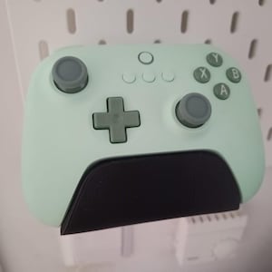 Puede incluir: Un mando de videojuegos inalámbrico verde menta con botones grises y un agarre negro. El mando tiene un panel direccional, botones de acción y joysticks. Está montado en una pared perforada blanca.