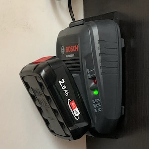 Op de afbeelding: Een zwarte Bosch AL 1830 CV batterijlader met een 2,5 Ah 18V batterij erin. De lader heeft een groen lampje dat aangeeft dat de batterij wordt opgeladen.