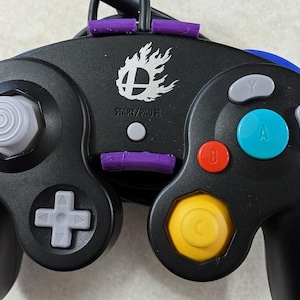 Puede incluir: Mando de videojuegos negro con una variedad de botones de colores, incluyendo rojo, azul y amarillo. El mando tiene un joystick y una cruceta grises. El texto "START/PAUSE" es visible, junto con un logotipo.