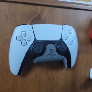 May include: A white and black PlayStation 5 DualSense controller with a grey controller stand. The controller has a symmetrical design with a touchpad and various buttons. The stand has the PlayStation logo.