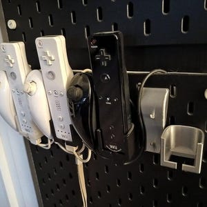 Puede incluir: Un panel perforado negro con varios mandos de Wii blancos y negros y una estación de carga. Los mandos están montados en el panel, con los mandos blancos a la izquierda y el mando negro a la derecha.