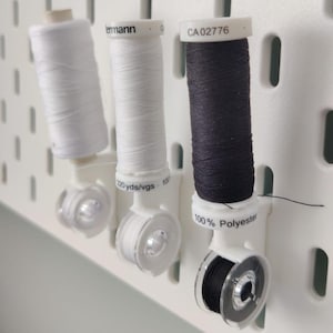 May include: Three spools of thread in white and black, mounted on a white pegboard. The spools are held in place by clear plastic holders. One spool is labeled "100% Polyester".